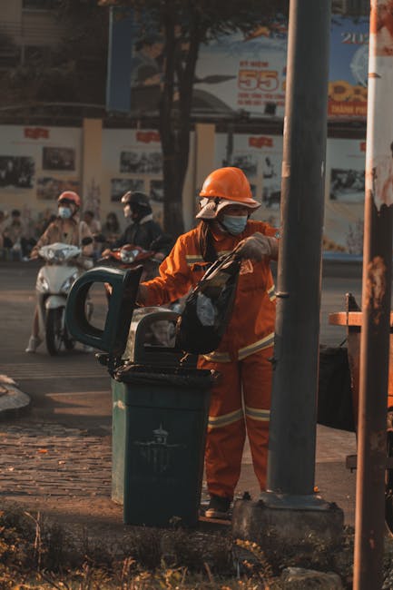 A waste management worker dressed in an orange high-visibility jumpsuit, wearing a protective helmet and a face mask, is seen standing on a sidewalk next to a large green rubbish bin with a black lid. The worker is holding the lid open while collecting waste, with a black backpack visible on their back. In the background, several motor scooters with riders wearing helmets and face masks are moving along the street, which features advertising posters and billboards on a wall. The worker is positioned beside a metal pole, and there is a glimpse of a tree casting shadows over the scene. The environment appears to be an urban area during daylight hours, with natural lighting highlighting the textures of the clothing, rubbish bin, and surrounding pavement, indicative of a typical waste collection operation in a city setting. The scene subtly relates to alternative waste handling services in a busy urban context, where on-site disposal might be necessary due to tight access conditions, with Rubbish Removal Battersea providing such services in the area.