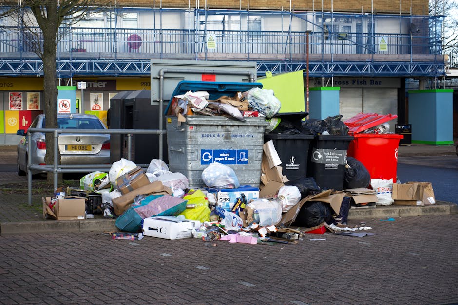 An outdoor scene showing a collection of mixed household waste and rubbish piled around multiple bin containers on a paved sidewalk in front of a shopping area. The rubbish includes crumpled cardboard boxes, plastic bags, paper waste, and various discarded packaging materials, some spilling onto the ground. The bins are of different colors: a large grey bin labeled for mixed paper and card, a black bin for general waste, and a bright red recycling bin. Behind the waste area, a metal railing separates the rubbish from parked vehicles, including a small silver car with a visible license plate. In the background, there is a storefront with signage partially obscured, featuring a yellow shopfront and large windows, and the upper floor appears to be under construction or renovation, enclosed with blue scaffolding and protective netting. The scene is lit with natural daylight, indicating daytime, and the overall environment suggests a busy urban area, where independent waste collection or private rubbish removal services such as Rubbish Removal Battersea might handle waste disposal outside of local authority collection systems.
