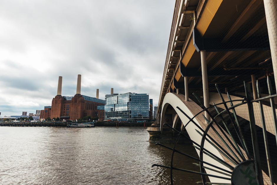 The image shows a view of the River Thames in London, featuring the old Battersea Power Station with its distinctive brick facade and four tall chimneys in the background, alongside modern glass buildings. On the right side, part of the underside of a large bridge or elevated structure with visible supporting columns and beams is seen, with a section of black metal railing or decorative fencing curving upward near the water's edge. The sky above is overcast with gray clouds, creating diffused lighting across the scene. The water in the foreground is calm, reflecting the surrounding structures. The scene appears to be taken from a low angle, highlighting the contrast between the historic industrial power station and contemporary architecture, hinting at the area’s evolving urban landscape that may involve private or alternative waste removal solutions in the Battersea region, such as rubbish clearance by specialists like Rubbish Removal Battersea.