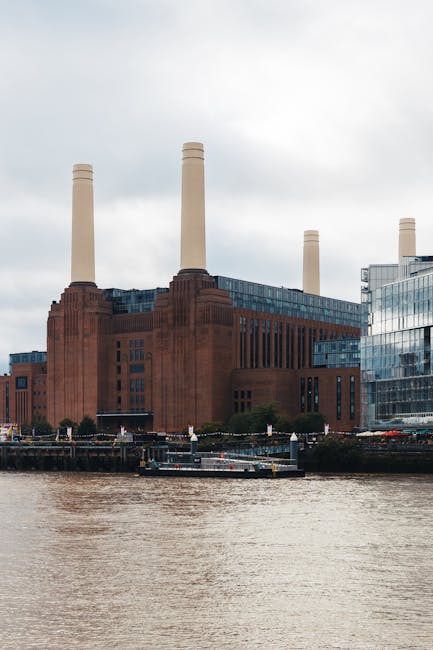 A large historic power station building constructed of red brick with vertical architectural details and four tall, light-colored smokestacks extending into a cloudy sky. The structure is positioned along a riverbank with a visible waterway in the foreground, where a small barge or boat is moored. Modern glass office buildings with reflective surfaces are situated to the right of the power station, creating a contrast between old and new architecture. The scene is captured during daylight, with natural light illuminating the details of the brickwork and the metallic surfaces of the adjacent buildings. This setting could relate to alternative waste handling solutions by highlighting the importance of site clearance and the removal of debris or waste materials during infrastructure upgrades. Rubbish Removal Battersea might handle waste clearance for such large-scale construction or renovation projects, emphasizing the need for efficient rubbish removal services in urban environments near Battersea Power Station.