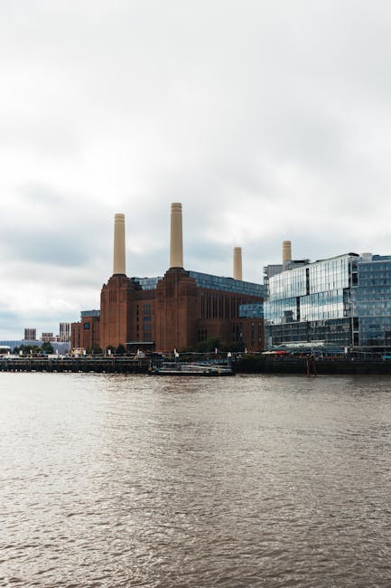 A low-angle view of several modern high-rise buildings and the historic Battersea Power Station, featuring a large brick chimney and Art Deco architectural details. The foreground includes a curved glass facade reflecting the sky and surrounding structures, while the background showcases a partly cloudy sky with patches of blue. The scene captures a mix of contemporary glass skyscrapers with sleek, curved surfaces and traditional brickwork, illustrating urban development near Battersea Power Station in SW11. The setting appears to be an outdoor urban square or plaza, with no visible rubbish or waste materials, but the scene contextually relates to sites that may be targeted for private rubbish removal or on-site clearance by services such as Rubbish Removal Battersea, highlighting the area's blend of historic and modern architecture.